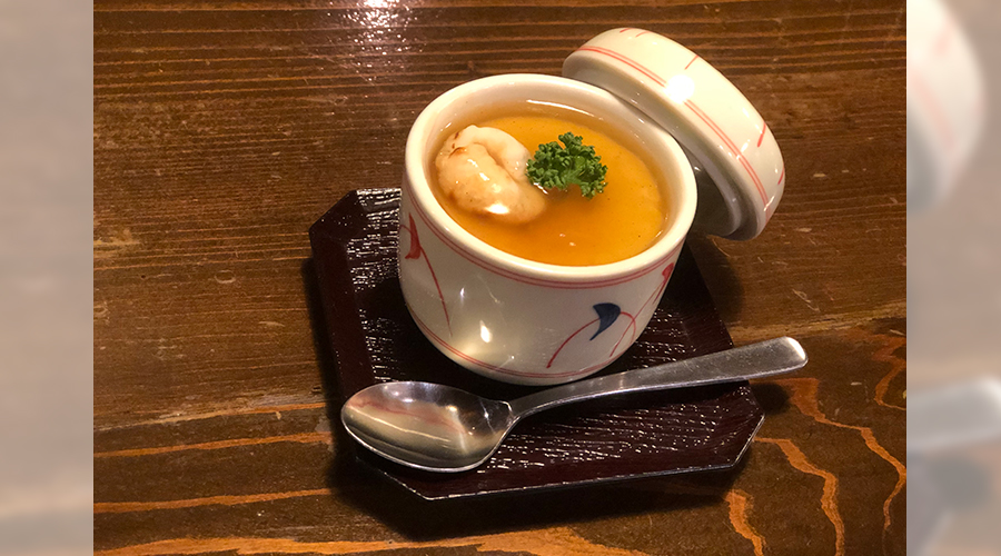 小さな陶器の器に入ったオレンジ色の茶碗蒸しにエビとパセリを添えた、暗いトレーに乗った一品