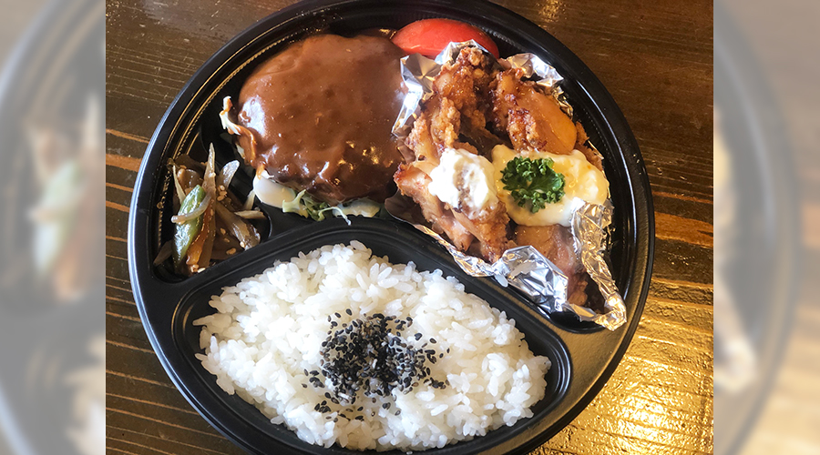 丸い弁当トレーに盛られたグレービーソースがかかったハンバーグと、揚げ物、ごま入りご飯、漬物
