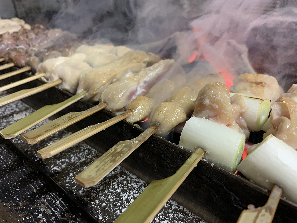 炭火の上で焼かれる串刺しの肉と長ねぎ、立ち上る煙のクローズアップ