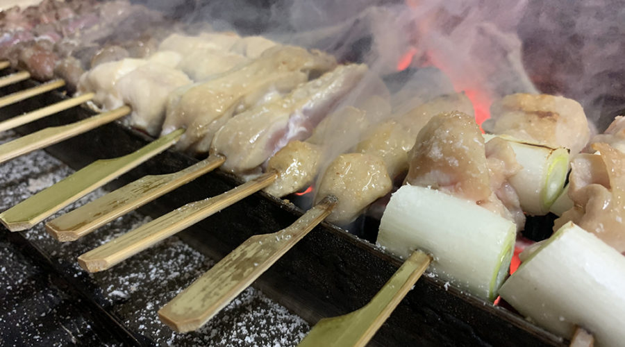 赤く燃える炭火の上で焼かれる串刺しの肉とねぎ、煙が立ち上る温かみのある雰囲気